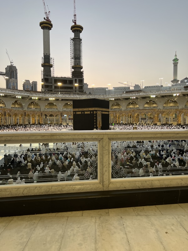A large crowd of people in white garments gathered around the Kaaba, the central black structure, in the courtyard of the Grand Mosque. Tall cranes and under-construction buildings are visible in the background, along with illuminated minarets and domes.