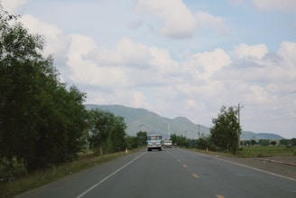 A rural road leading towards distant hills under a partly cloudy sky. Several vehicles are visible, including a bus and a car, moving along the road. Trees line the sides, and power lines run parallel to the road.