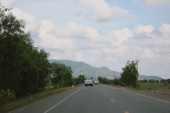 A rural road leading towards distant hills under a partly cloudy sky. Several vehicles are visible, including a bus and a car, moving along the road. Trees line the sides, and power lines run parallel to the road.