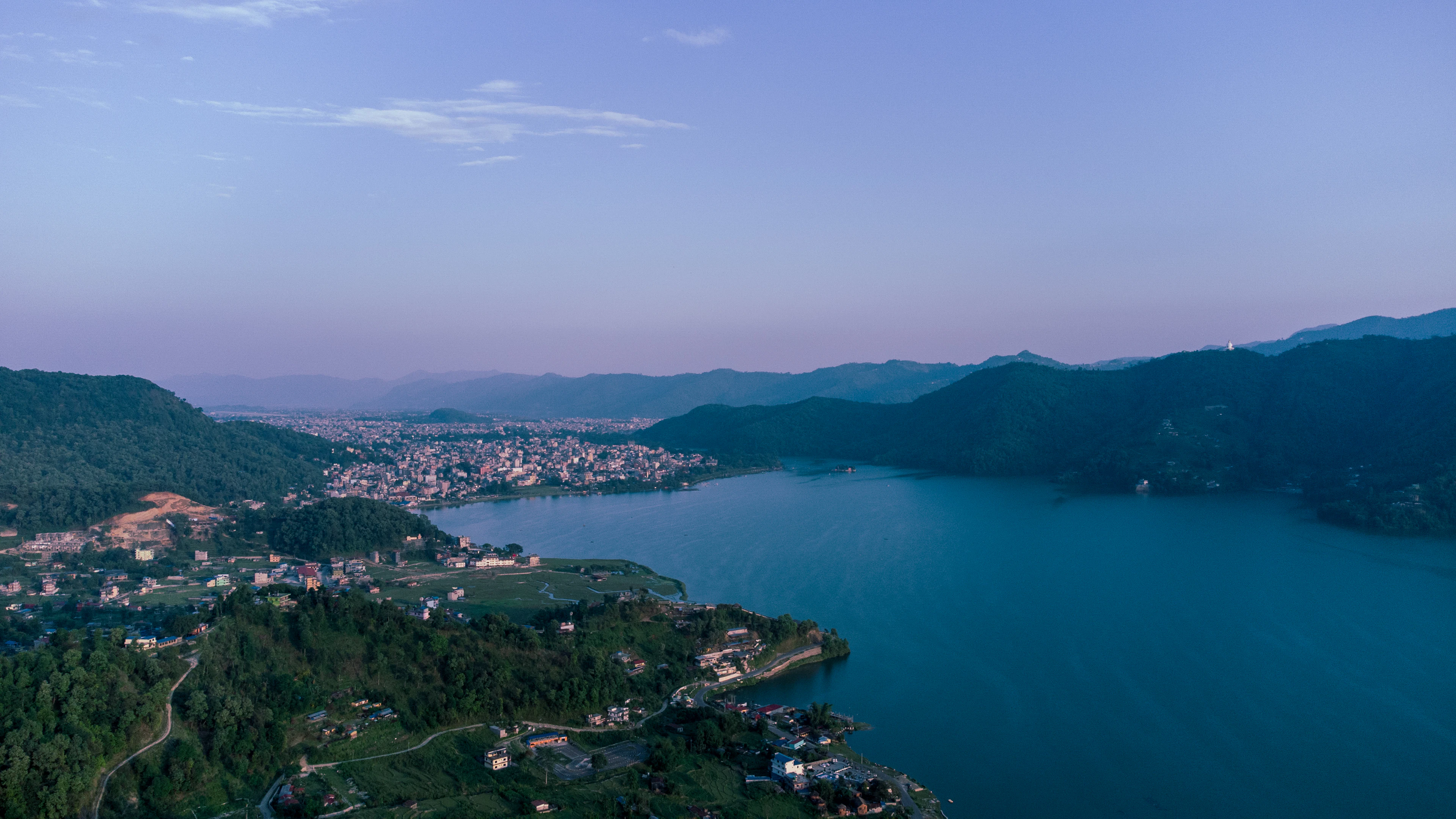 Phewa Lake at dusk from the air, its still blue water curving between forested ridges with the town of Pokhara spread across the valley floor beyond under a violet sky.