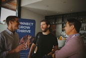 Three individuals are engaged in conversation in a bar or cafe setting. One person is gesturing with their hand while the others smile, suggesting a positive interaction. Behind them, a banner displays the words 'Connect, Grow, Thrive.'.