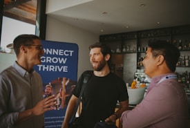 Three individuals are engaged in conversation in a bar or cafe setting. One person is gesturing with their hand while the others smile, suggesting a positive interaction. Behind them, a banner displays the words 'Connect, Grow, Thrive.'.