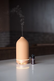 A beige-colored essential oil diffuser is placed on a glossy surface with a small bottle of essential oil next to it. The diffuser is emitting a trail of vapor, suggesting it is in use. The background consists of a dark, dimly lit area.