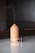 A beige-colored essential oil diffuser is placed on a glossy surface with a small bottle of essential oil next to it. The diffuser is emitting a trail of vapor, suggesting it is in use. The background consists of a dark, dimly lit area.