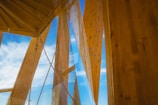 Wooden architectural beams and panels stretch upwards towards a bright blue sky with scattered clouds. Transparent glass elements intersect with the wooden structure, adding a modern touch.