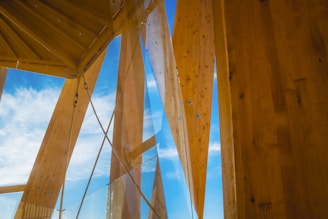 Wooden architectural beams and panels stretch upwards towards a bright blue sky with scattered clouds. Transparent glass elements intersect with the wooden structure, adding a modern touch.