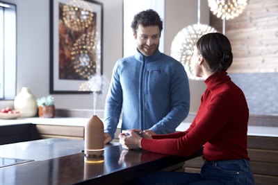 A man and woman are in a modern kitchen. The man, wearing a blue fleece, is standing and smiling at the woman, who is seated and wearing a red sweater. They are engaged in conversation with a diffuser emitting vapor on the table between them. The kitchen is decorated with warm lighting and contemporary artwork.