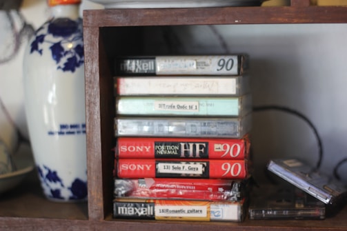 Several audio cassette tapes are stacked vertically on a wooden shelf. The tapes have various labels, some with handwritten text. A decorative vase with blue floral patterns is partially visible to the left.