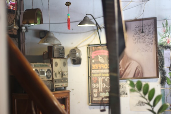 A vintage indoor setting featuring various retro objects. There is a large, old-fashioned radio and stereo reel-to-reel recorder placed on a wooden cabinet. A poster advertising coffee hangs on the wall, alongside a faded photograph or painting, partially occluded by a plant with broad leaves. Industrial-style pendant lamps hang from the ceiling, and a red tassel ornament is suspended from a wire. The space has a cluttered yet nostalgic ambiance, enhanced by wires and pipes visible throughout.