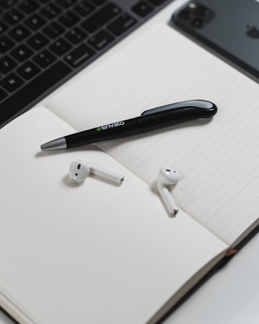 A black pen with 'envato' printed on it rests on an open lined notebook. Two wireless earbuds are placed nearby on the notebook. The corner of a laptop keyboard and a smartphone are partially visible, creating a workspace setting.
