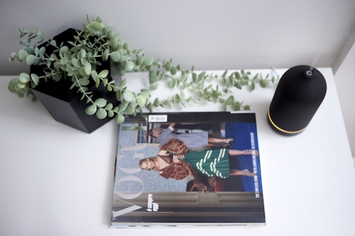 A magazine is placed on a white surface next to a black potted plant with green foliage and a black cylindrical object with a yellow stripe. The magazine features a fashion photograph on the cover.