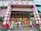 A storefront with red and white signage featuring Chinese characters. The store has an awning and displays a variety of goods such as dried foods, snacks, and other items. Several people, some wearing masks, are walking by or standing in front of the shop.