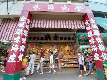 A storefront with red and white signage featuring Chinese characters. The store has an awning and displays a variety of goods such as dried foods, snacks, and other items. Several people, some wearing masks, are walking by or standing in front of the shop.