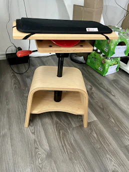 A wooden stool-like structure with a padded seat on top. The stool has a natural wood finish with round and curved edges. There's a red lever on one side, likely for adjusting the height or position. In the background, there are green packages of bottled drinks stacked neatly, and a cardboard box on a wooden floor.