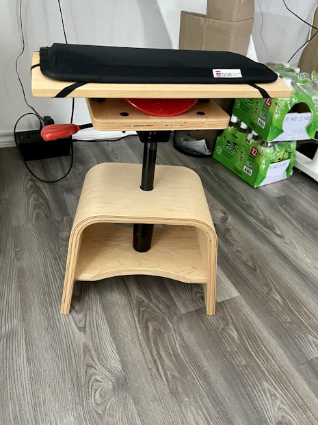 A wooden stool-like structure with a padded seat on top. The stool has a natural wood finish with round and curved edges. There's a red lever on one side, likely for adjusting the height or position. In the background, there are green packages of bottled drinks stacked neatly, and a cardboard box on a wooden floor.