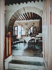 A cozy, rustic interior with stone arches and wooden beams. There is a dining area with a table covered by a patterned tablecloth and surrounded by chairs. The space is warmly lit by natural light streaming through the large windows.