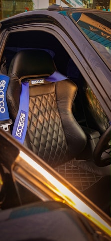 A luxurious car interior features a black leather racing seat with a diamond-quilted pattern. The seat is equipped with blue Sparco branded racing harnesses. The setting appears to be well-lit, highlighting the elegance and sporty design.