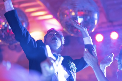 A man dressed in a suit appears to be dancing energetically at a party under disco balls and colorful lights. His arms are raised, and he looks engaged with the music and environment. Other people are partially visible in the background, also appearing to enjoy the lively atmosphere.