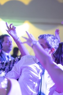 A group of people are dancing energetically at a social event. The scene is lively with bright, colorful lighting casting hues of purples and yellows across the room. The individuals are dressed formally, with one person wearing a white shirt and a tie, and others around them seem to be in motion with raised arms and blurred movements.