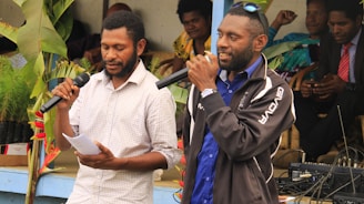 Two men are standing and speaking into microphones. One man, dressed in a checkered shirt, holds paper in his hand, while the other wears a blue shirt and a dark jacket. Several people are seated in the background, among green plants and some audio equipment on a table.