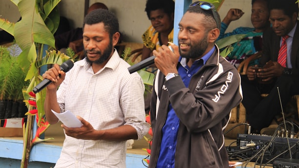 Two men are standing and speaking into microphones. One man, dressed in a checkered shirt, holds paper in his hand, while the other wears a blue shirt and a dark jacket. Several people are seated in the background, among green plants and some audio equipment on a table.