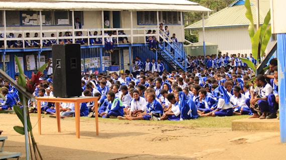 A large group of students seated outdoors in a courtyard area, all wearing matching blue and white uniforms. A small speaker is placed on a table towards the front. The background shows a raised building with visible classrooms and balconies, where more students can be seen observing the event. Lush green plants are scattered around the area.