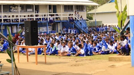 A large group of students seated outdoors in a courtyard area, all wearing matching blue and white uniforms. A small speaker is placed on a table towards the front. The background shows a raised building with visible classrooms and balconies, where more students can be seen observing the event. Lush green plants are scattered around the area.