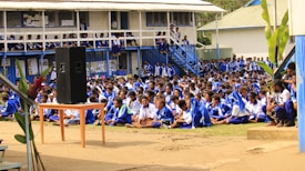 A large group of students seated outdoors in a courtyard area, all wearing matching blue and white uniforms. A small speaker is placed on a table towards the front. The background shows a raised building with visible classrooms and balconies, where more students can be seen observing the event. Lush green plants are scattered around the area.