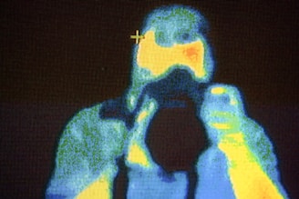 A thermal image captured using infrared technology, showing a person or object with varying heat signatures. Warmer areas appear in yellow and red tones, while cooler areas are depicted in blue and green tones. The image includes a crosshair, possibly indicating a point of focus or measurement.