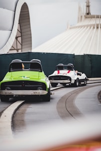 Two small cars on a track with a futuristic architectural background. The cars are brightly colored, one green and one white with red stripes, and appear to be part of an amusement park ride. A section of a large, dome-shaped building is visible in the background.