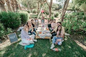 A group of people sitting on the grass in a park setting, gathered around a low picnic table set with plates and drinks. They appear to be enjoying a casual outdoor meal. The surrounding area is lush with green foliage and trees. A signboard with text is visible on the side.