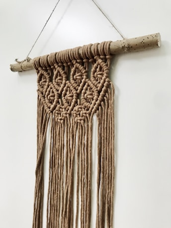 A handcrafted macrame wall hanging is suspended from a natural wooden rod. The intricate pattern includes geometric shapes created by knotted brown cord, which extends downward into long fringes. The neutral tones and simple design contribute to a rustic and earthy aesthetic.