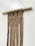 A handcrafted macrame wall hanging is suspended from a natural wooden rod. The intricate pattern includes geometric shapes created by knotted brown cord, which extends downward into long fringes. The neutral tones and simple design contribute to a rustic and earthy aesthetic.