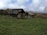 Two rustic, old wooden cottages with stone and tin roofs are set on a grassy and slightly uneven landscape. The area is surrounded by trees with autumn foliage under a cloudy sky, creating a serene rural scene.