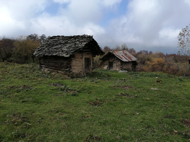 Two rustic, old wooden cottages with stone and tin roofs are set on a grassy and slightly uneven landscape. The area is surrounded by trees with autumn foliage under a cloudy sky, creating a serene rural scene.