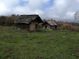 Two rustic, old wooden cottages with stone and tin roofs are set on a grassy and slightly uneven landscape. The area is surrounded by trees with autumn foliage under a cloudy sky, creating a serene rural scene.
