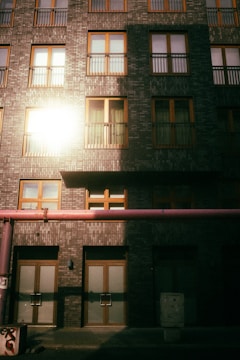 A multi-story building facade with uniformly arranged windows framed in wood against a textured brick surface. Sunlight reflects off one of the windows, casting a bright glare. A pink pipe runs horizontally across the lower part of the structure, adding a contrasting element to the scene.