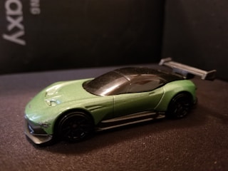 A green toy car with a sleek design and a black roof is positioned on a dark surface. The car features a prominent rear wing and dark wheels, emphasizing its sporty appeal.