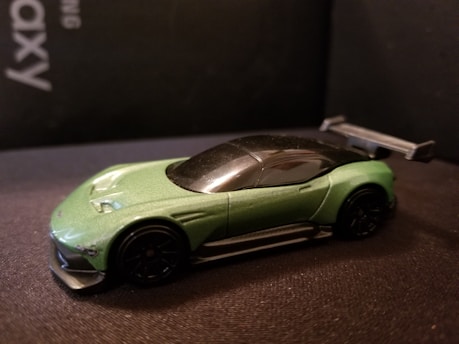 A green toy car with a sleek design and a black roof is positioned on a dark surface. The car features a prominent rear wing and dark wheels, emphasizing its sporty appeal.