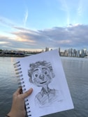 A hand holds a sketchpad with a detailed pencil drawing of an animated character. The background features a city skyline across a body of water under a partly cloudy sky. The scene captures the horizon with a gentle transition from water to high-rise buildings and then to the sky.