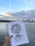 A hand holds a sketchpad with a detailed pencil drawing of an animated character. The background features a city skyline across a body of water under a partly cloudy sky. The scene captures the horizon with a gentle transition from water to high-rise buildings and then to the sky.
