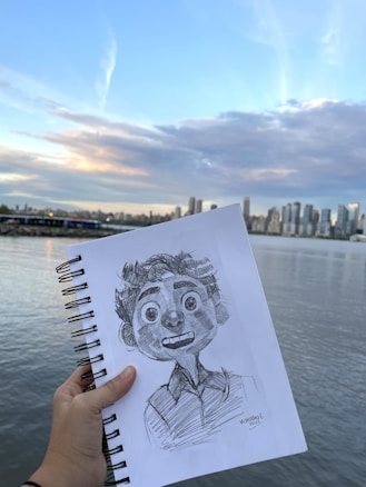 A hand holds a sketchpad with a detailed pencil drawing of an animated character. The background features a city skyline across a body of water under a partly cloudy sky. The scene captures the horizon with a gentle transition from water to high-rise buildings and then to the sky.