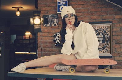 A person wearing a white hoodie and beanie is sitting on a table with one leg extended. They are holding a popsicle and wearing glasses, looking pensively sideways. A skateboard rests beside them on the table. The setting appears cozy with brick walls and warm lighting, featuring decorative signs and a tranquil atmosphere.