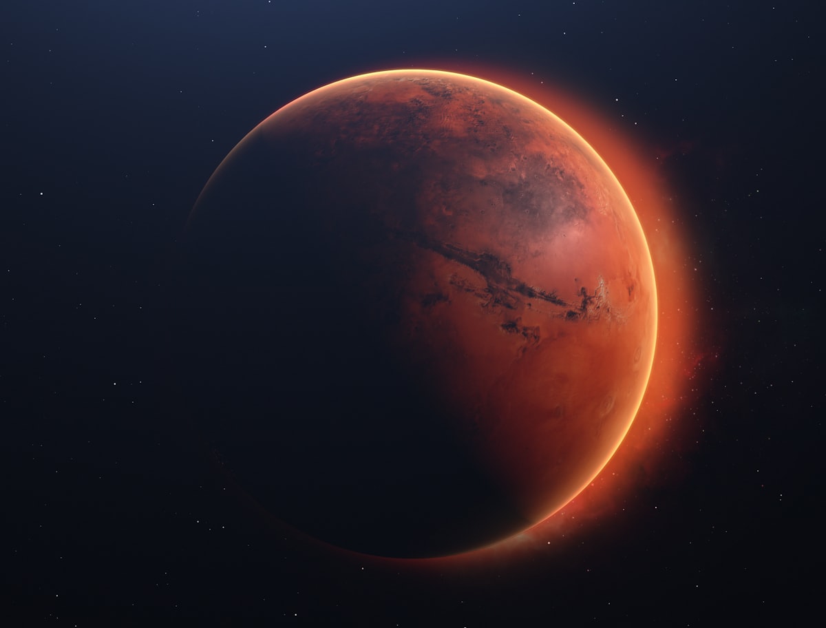 A reddish, cratered planet hangs against deep black space, sunlight catching one limb so the rest sits in shadow.