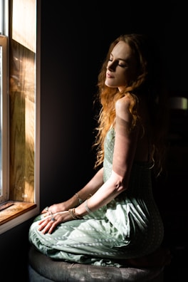A woman with long, wavy hair is sitting by a window, bathed in natural sunlight. She is wearing a green dress and various rings and bracelets. Her eyes are closed, and she appears to be in a calm or meditative state. The background is dark, contrasting with the bright light coming from the window.