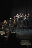 A group of musicians dressed in black formal wear perform on stage with various wind instruments, such as trumpets, trombones, and saxophones. They are arranged in rows, and some are seated while others stand. The background is a dark curtain, and a piano is visible in the foreground.