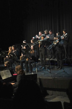 A group of musicians dressed in black formal wear perform on stage with various wind instruments, such as trumpets, trombones, and saxophones. They are arranged in rows, and some are seated while others stand. The background is a dark curtain, and a piano is visible in the foreground.