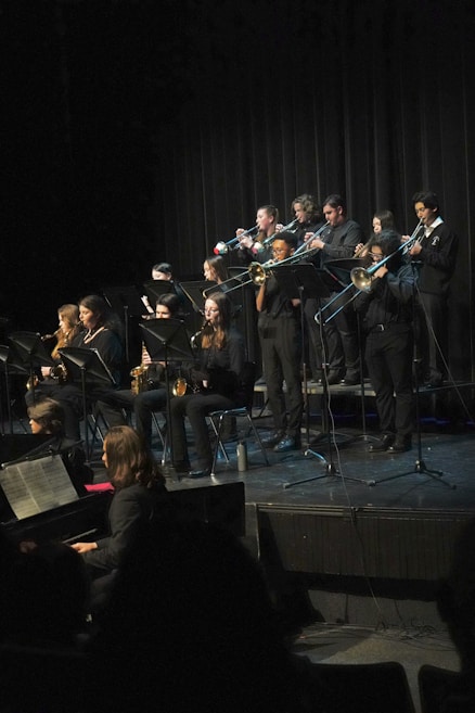 A group of musicians dressed in black formal wear perform on stage with various wind instruments, such as trumpets, trombones, and saxophones. They are arranged in rows, and some are seated while others stand. The background is a dark curtain, and a piano is visible in the foreground.