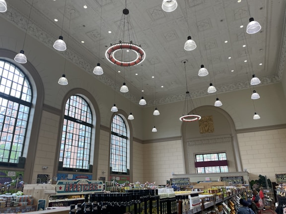 A spacious indoor market with high ceilings, featuring large arched windows and elaborate ceiling decorations. Numerous hanging lights illuminate the area. Below, rows of shelves with various items like bottles and food products are visible. People are browsing the aisles, contributing to the bustling atmosphere.