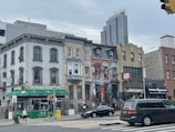 A city street with a row of diverse architectural buildings including a halal Chinese food restaurant, colorful murals on one building, and a deli. Pedestrians are walking on the sidewalk, and a few vehicles are visible on the street under a cloudy sky.